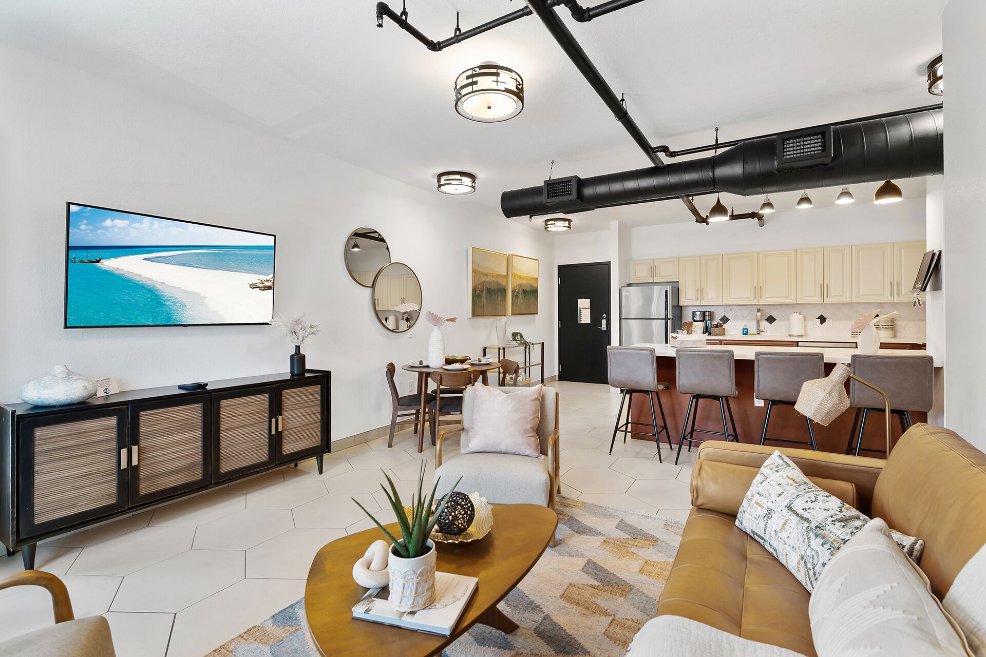 A modern living room with a large flat-screen TV, a coffee table with a plant, a sofa, and a dining area with a kitchen in the background.