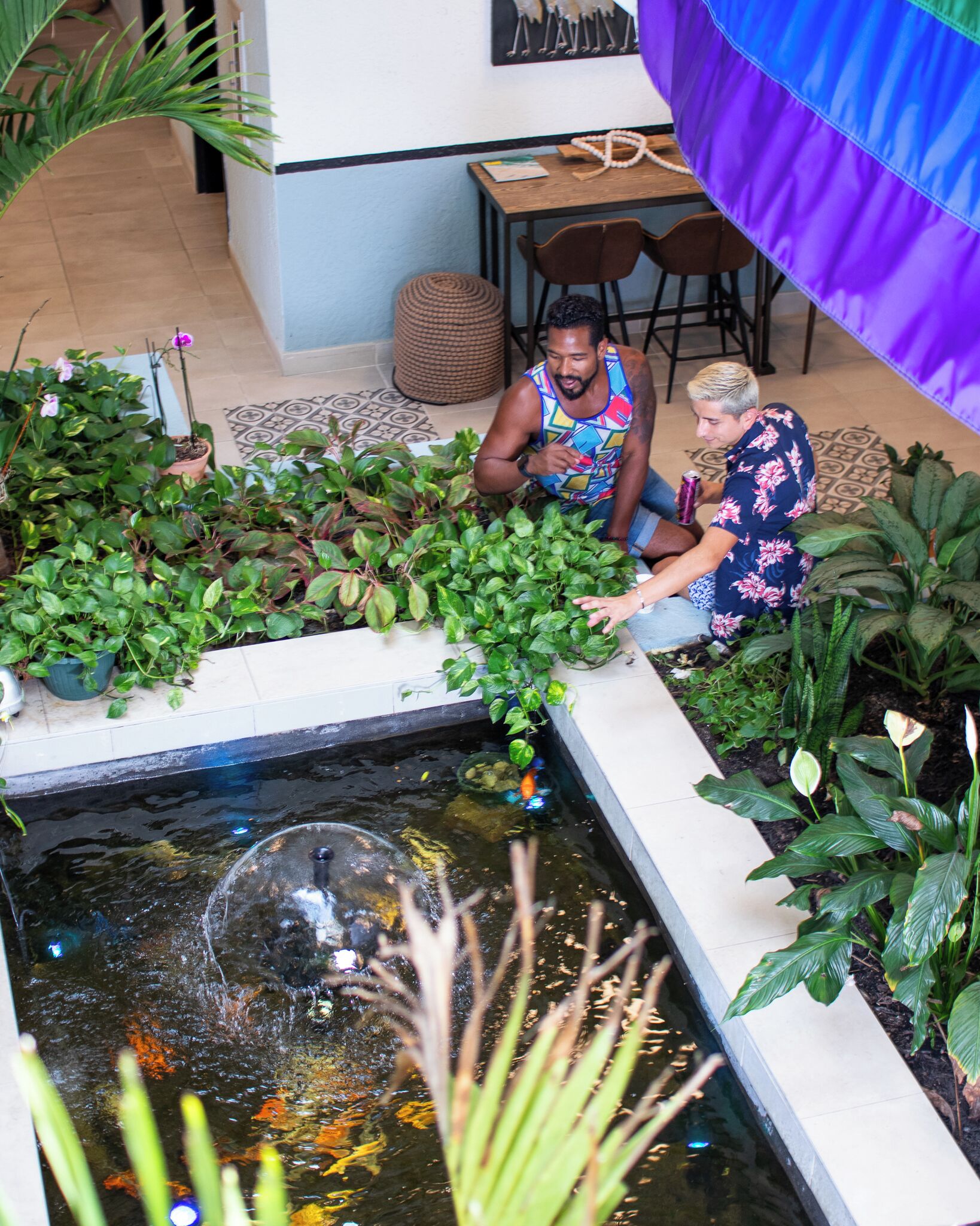 A small indoor garden with a pond, potted plants, and two people sitting by the water.