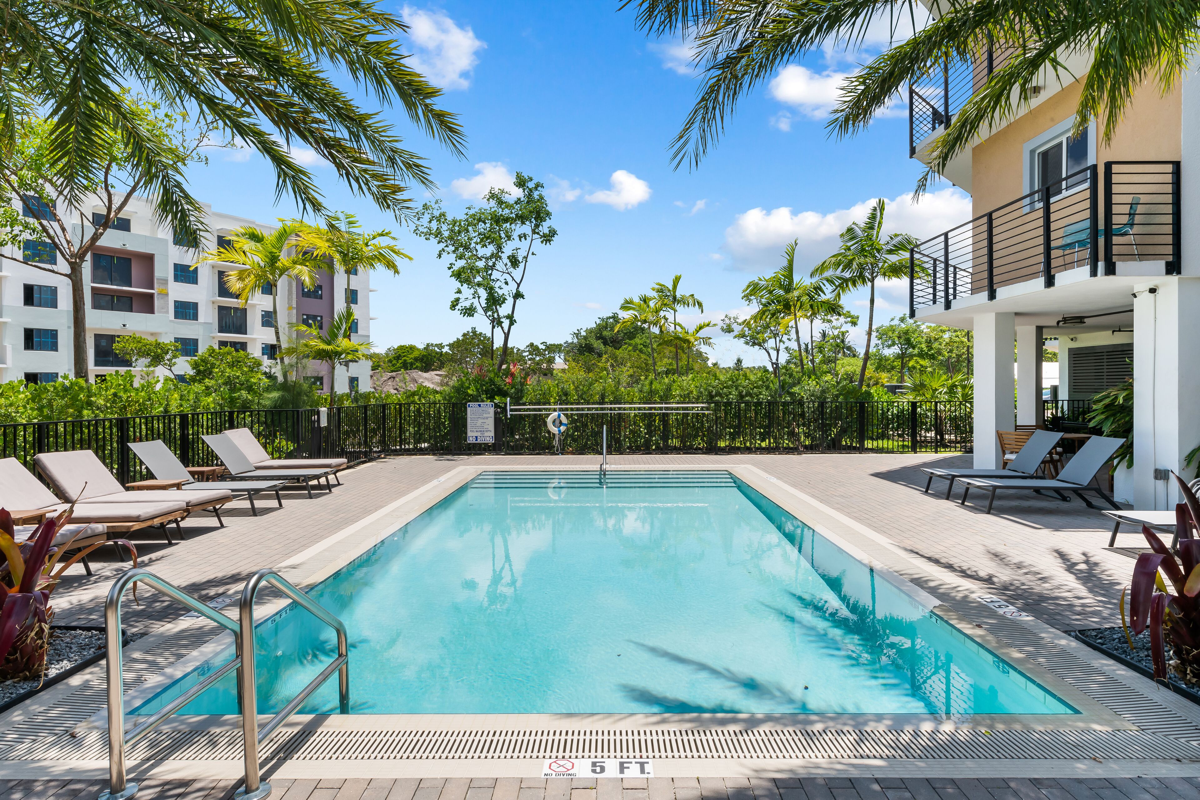 A swimming pool with lounge chairs, surrounded by palm trees and a building with balconies.