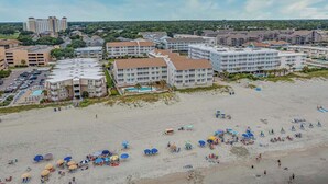 Ein Hotel direkt am Strand mit mehreren Gebäuden, einem Parkplatz und einer Sandstrandfläche mit Sonnenschirmen.