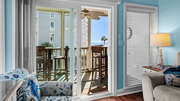 Ein Zimmer mit einer Glasschiebetür, die auf einen Balkon mit Meerblick führt. Es gibt ein Sofa mit gemustertem Bezug und eine Lampe auf einem Beistelltisch.