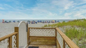 Eine Holzpromenade führt zu einem Strand mit Sonnenschirmen und Menschen in der Ferne.