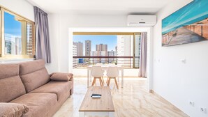 Una sala moderna con sofá, una mesa de centro y un comedor con sillas. Hay un ventanal con vistas a edificios y un cuadro en la pared.