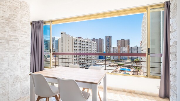 Un balcón con mesa y sillas, que ofrece vistas de un paisaje urbano con edificios y una piscina.