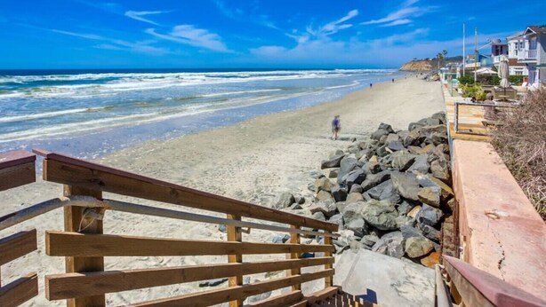Una spiaggia con una scalinata di legno che scende verso la riva, una barriera rocciosa e case sullo sfondo.