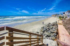 Une plage avec un escalier en bois descendant jusqu’au rivage, une barrière rocheuse et des maisons en arrière-plan.