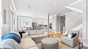A modern living room with a sectional sofa, ottoman, and a chair. A kitchen with white cabinets and stainless steel appliances is visible in the background. - Spacious Seagrove Escape Cart Bikes Pool Near Beach (Santa Rosa Beach)