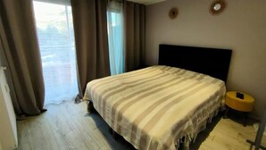Ein Schlafzimmer mit einem großen Bett, einem kleinen runden Tisch und Blick ins Freie durch ein Fenster mit Vorhängen.