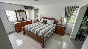 Ein Schlafzimmer mit einem großen Bett, Holznachttischen, einer Kommode, einem Spiegel und zwei mit Vorhängen versehenen Fenstern.
