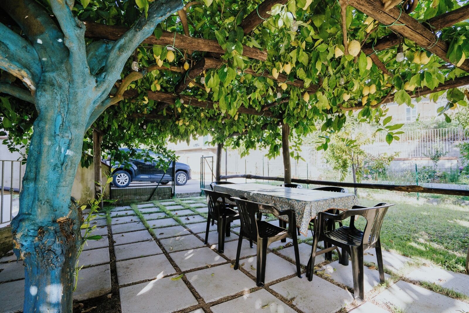 Eine Terrasse im Freien mit Tisch und Stühlen, ein geparktes Auto im Hintergrund und ein Baum mit grünen Blättern.