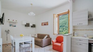 En kompakt opholdsstue med spiseplads, sofa, rød lænestol, køkken med kaffemaskine og vindue med udsigt til et skovområde.