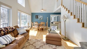 A spacious living room with a high ceiling, wooden beams, a staircase, and large windows.