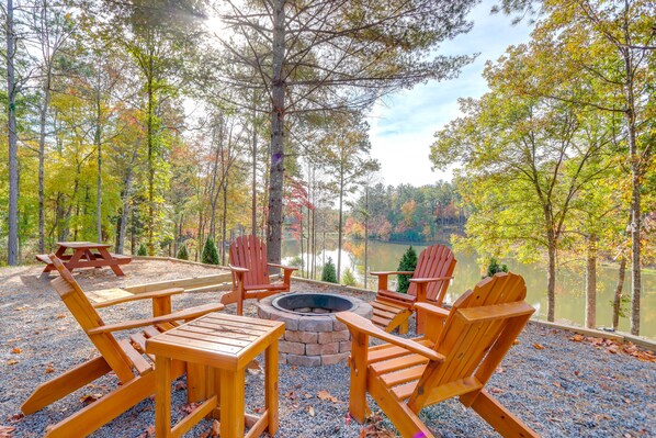 Una terraza de madera con sillas Adirondack y un hogar de piedra, con vista a un lago rodeado de árboles.