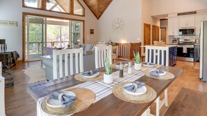Ein geräumiger Essbereich mit hoher Decke und großen Fenstern, ein Holztisch gedeckt mit Platzsetzern und Mittelstücken, und Blick auf die Küche sowie die Terrassenfläche.