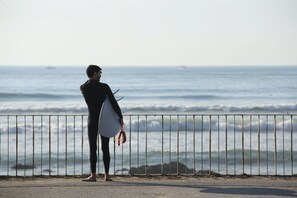 A person in a wetsuit holding a surfboard, standing by a railing overlooking the ocean.