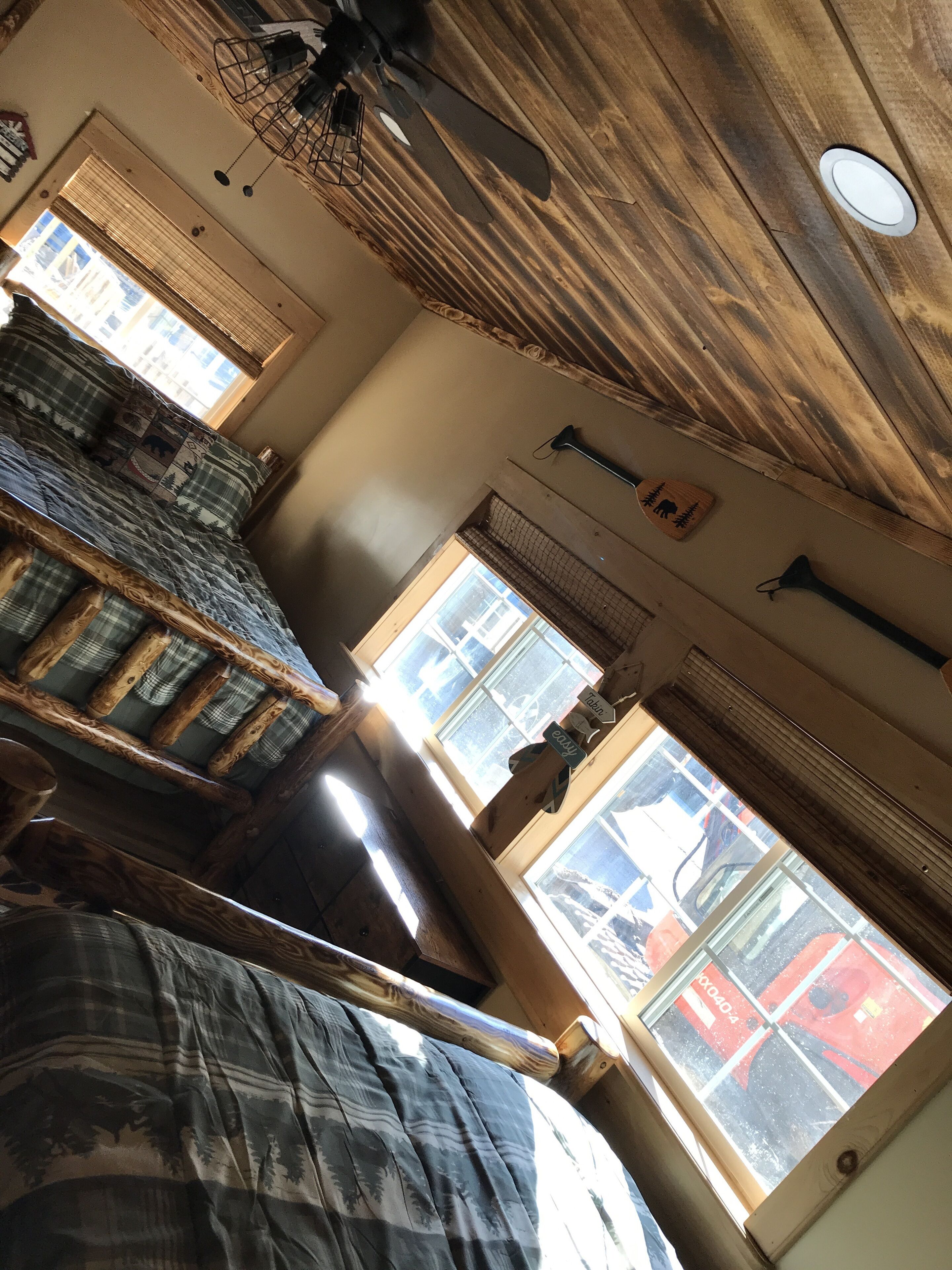 A rustic room with wooden beams, a bed with a plaid blanket, and a window with a view of a red vehicle outside.