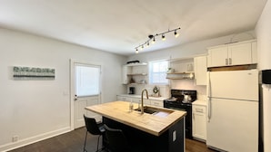 A compact kitchen with a central island, a stove, a refrigerator, and a window with blinds. - Point on Pollock - Brewery Row - Brim of Downtown (New Bern)
