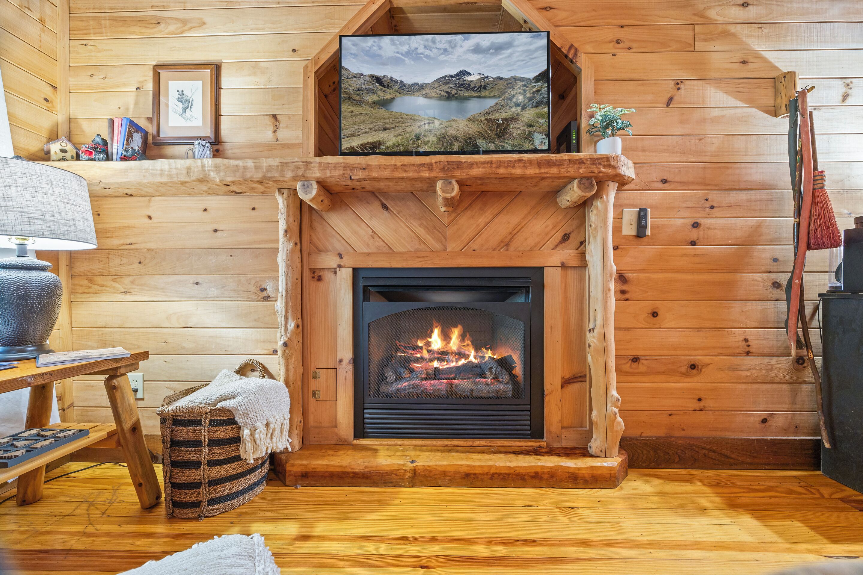 A cozy cabin interior with a fireplace, wooden walls, a television mounted above it, and a small table with a lamp.