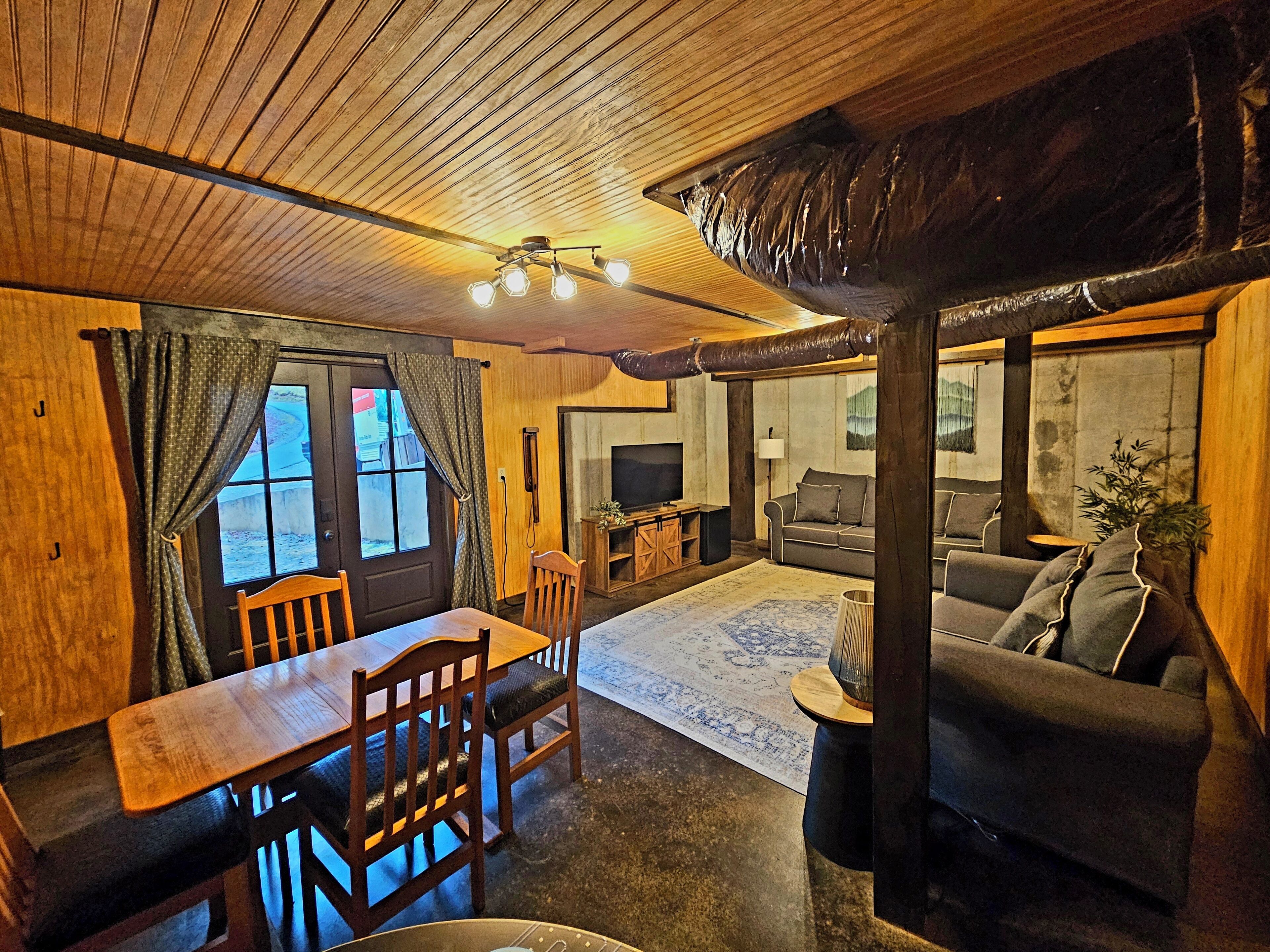 A cozy living room with a wooden ceiling, a dining area with a table and chairs, a sofa, and a television.