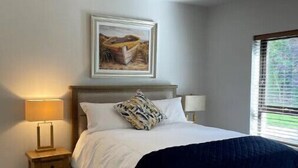 A bedroom with a bed, a nightstand, a lamp, a ceiling light, a painting, and a window.