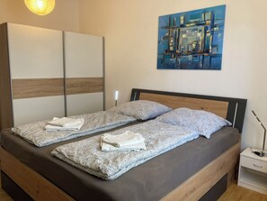 Uma cama de casal com roupa de cama estampada e duas almofadas, uma mesa de cabeceira com abajur e um guarda-roupa com portas espelhadas.