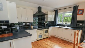 A kitchen with white cabinets, a black tiled backsplash, wooden flooring, and a window with curtains. - Ty Mawr (Haverfordwest)