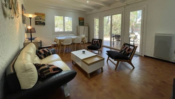 Un salón espacioso con un sofá blanco, una mesita de centro y una silla de madera. Se aprecia una zona de comedor con mesa y sillas al fondo.