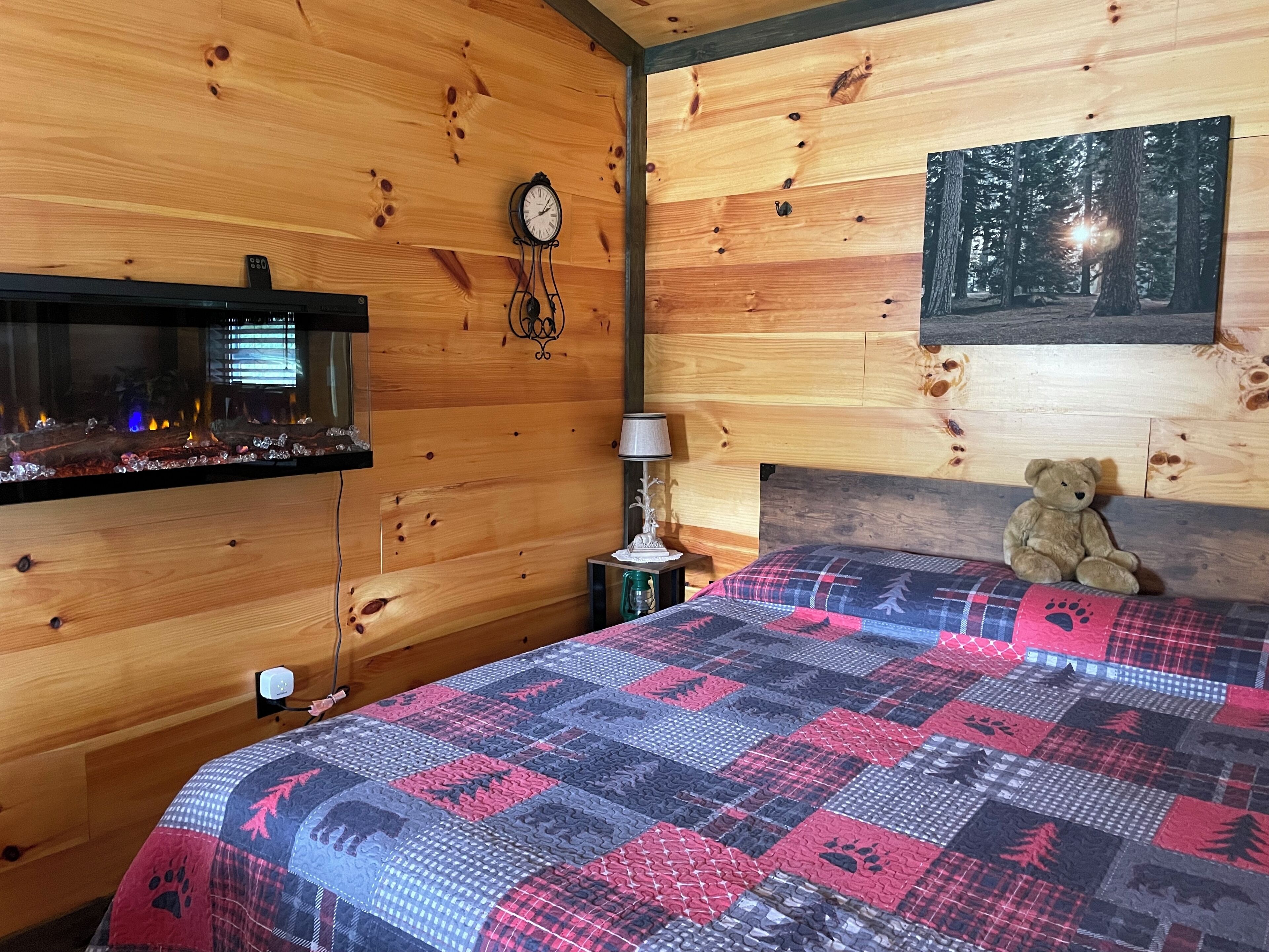 Una habitació acollidora amb una paret de fusta, un llit amb un estenedora estampada, un os de peluix, un rellotge i un quadre d'un bosc.