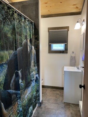 A bathroom with a shower curtain depicting a forest scene, a window with blinds, a sink, and a towel rack.