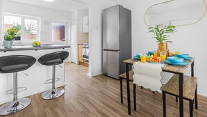 A modern kitchen with a refrigerator, two bar stools, a dining table with blue bowls and fruits, and a view of the outside through a window. - 3 Bed House Sleeps 8 -near Heathrow -free Parking (Hounslow)