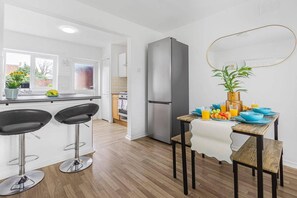 Una cocina moderna con refrigerador, dos taburetes de bar, una mesa de comedor con cuencos azules y frutas, y vistas al exterior a través de una ventana.