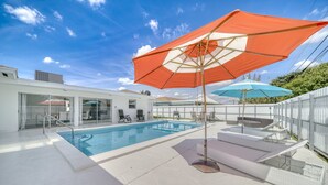 En swimmingpool med ett vitt poolområde, en loungehörna med ett vitt staket och ett stort parasoll som ger skugga.