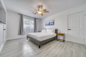 Una camera da letto moderna con un letto grigio, pareti bianche, una ventola a soffitto, una finestra con tende e una porta.
