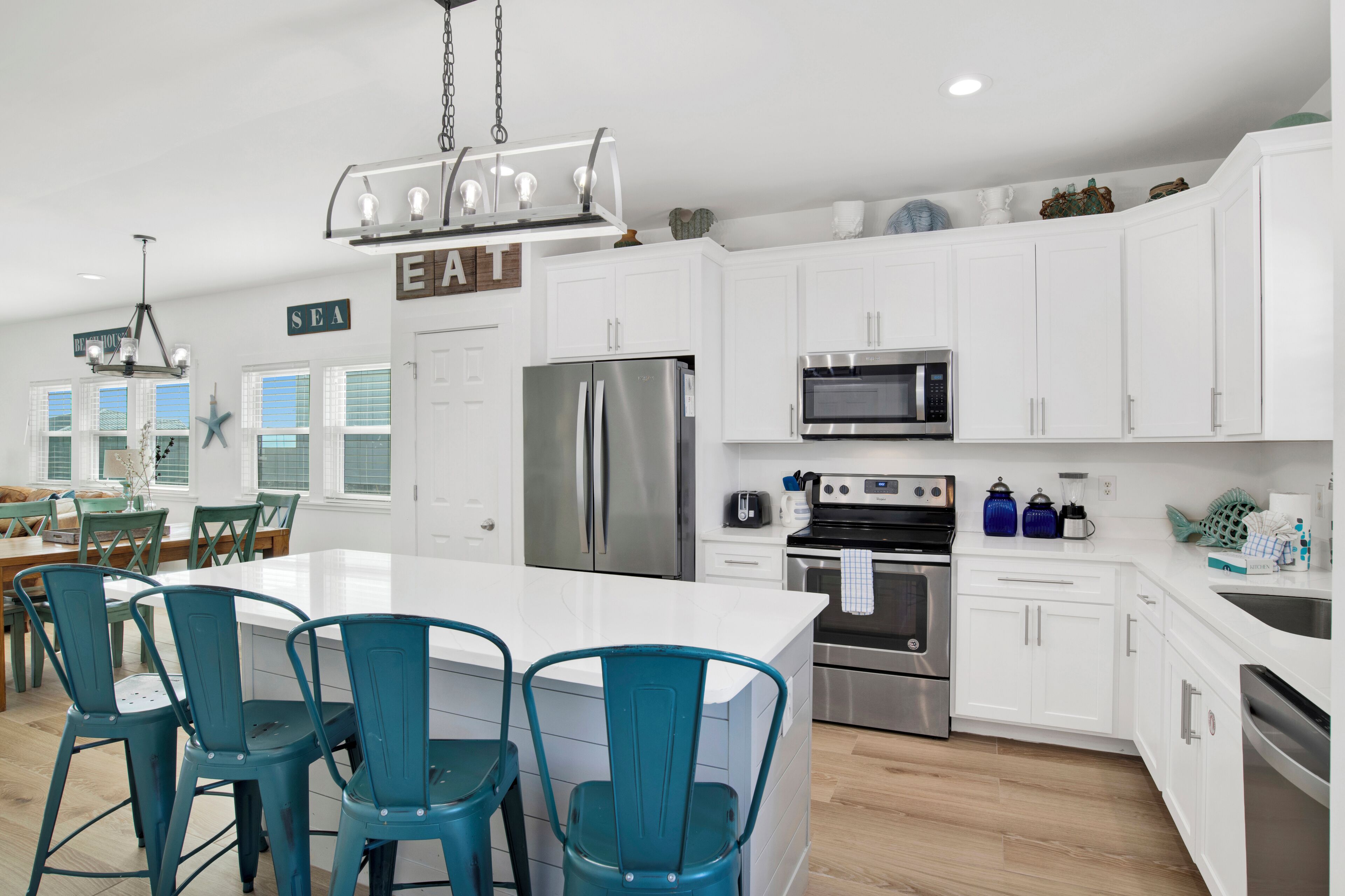 Una cocina moderna con armarios blancos, electrodomésticos de acero inoxidable, sillas color turquesa y una isla central.