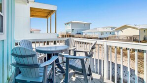 Una terraza de madera con sillas Adirondack y una mesa, con vista a una playa y casas de playa.