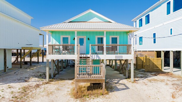 Una casa de playa de dos pisos con balcón, terraza de madera y zona de playa arenosa.