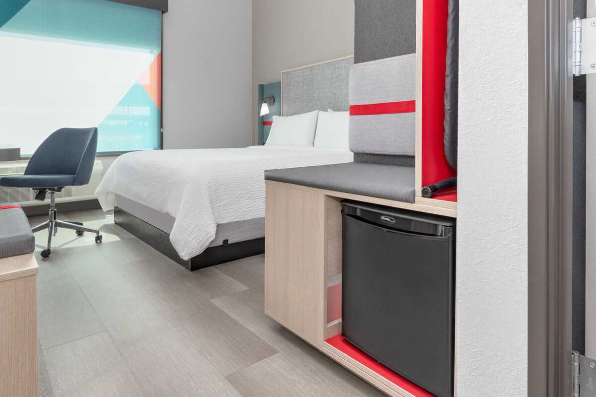 A modern hotel room with a bed, a desk, a chair, and a compact kitchenette.