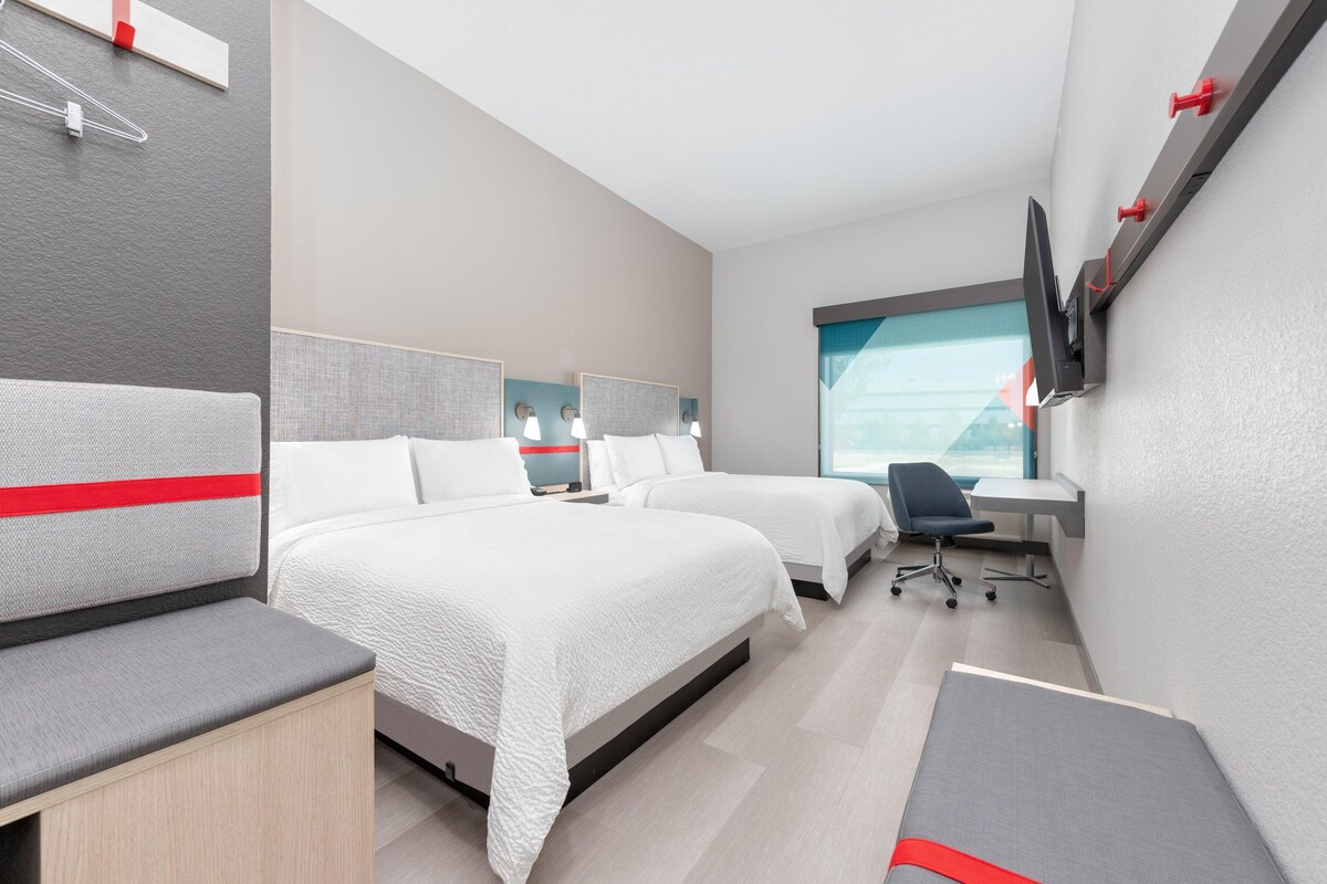 A modern hotel room with two beds, a desk, a chair, and a TV mounted on the wall.