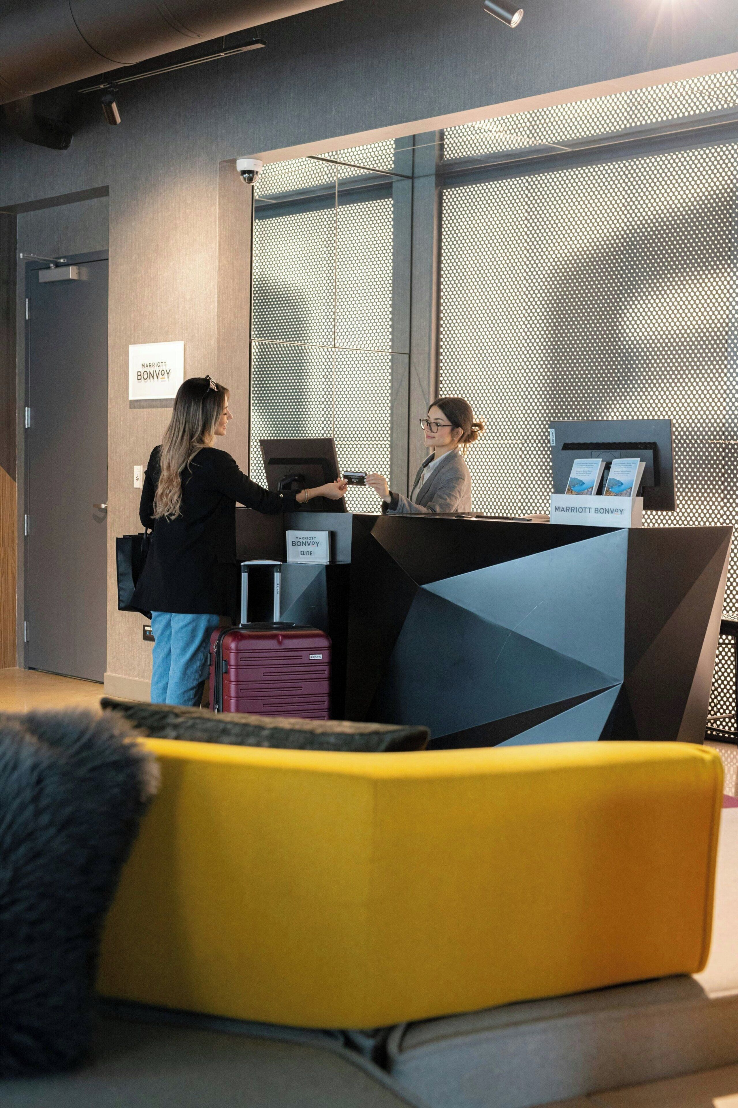 Un área de recepción de diseño moderno, con mostrador de registro, una zona de estar con una silla amarilla y un cartel visible del hotel Marriott Bonvoy.