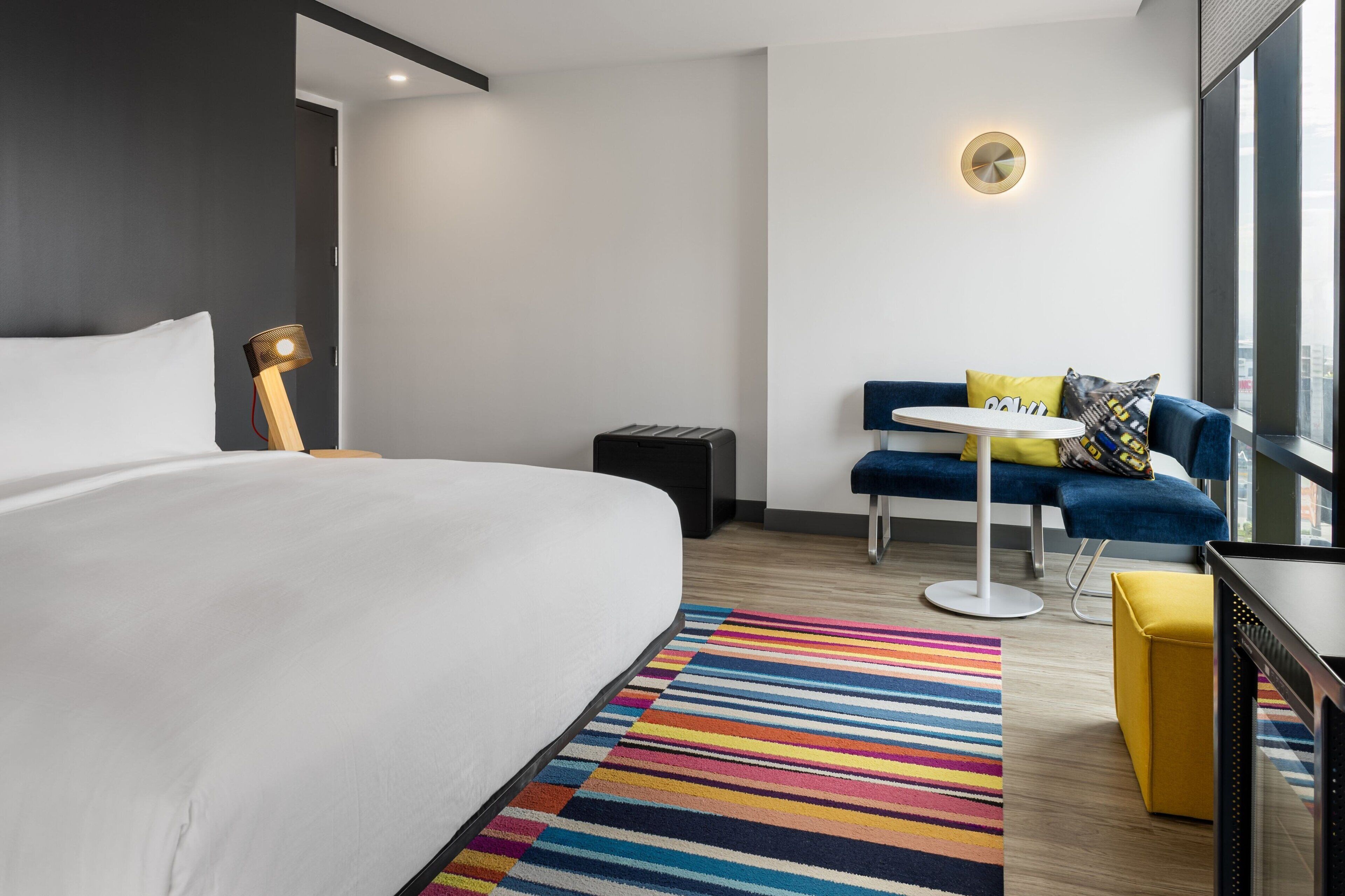A modern hotel room with a large bed, a small table, a sofa, and a colorful striped rug.