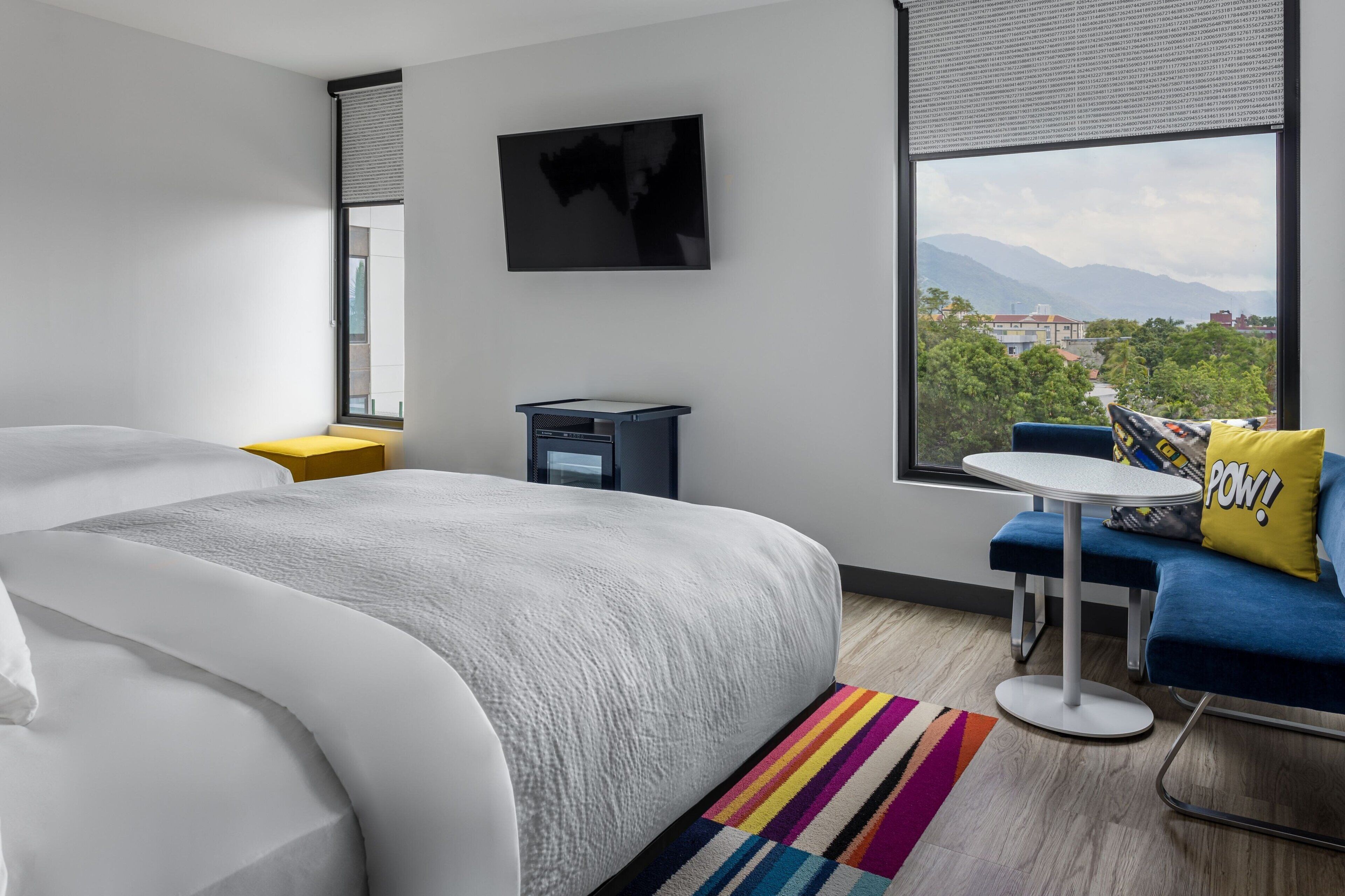 A modern hotel room with a large bed, a small desk, a TV, and a view of greenery and mountains.