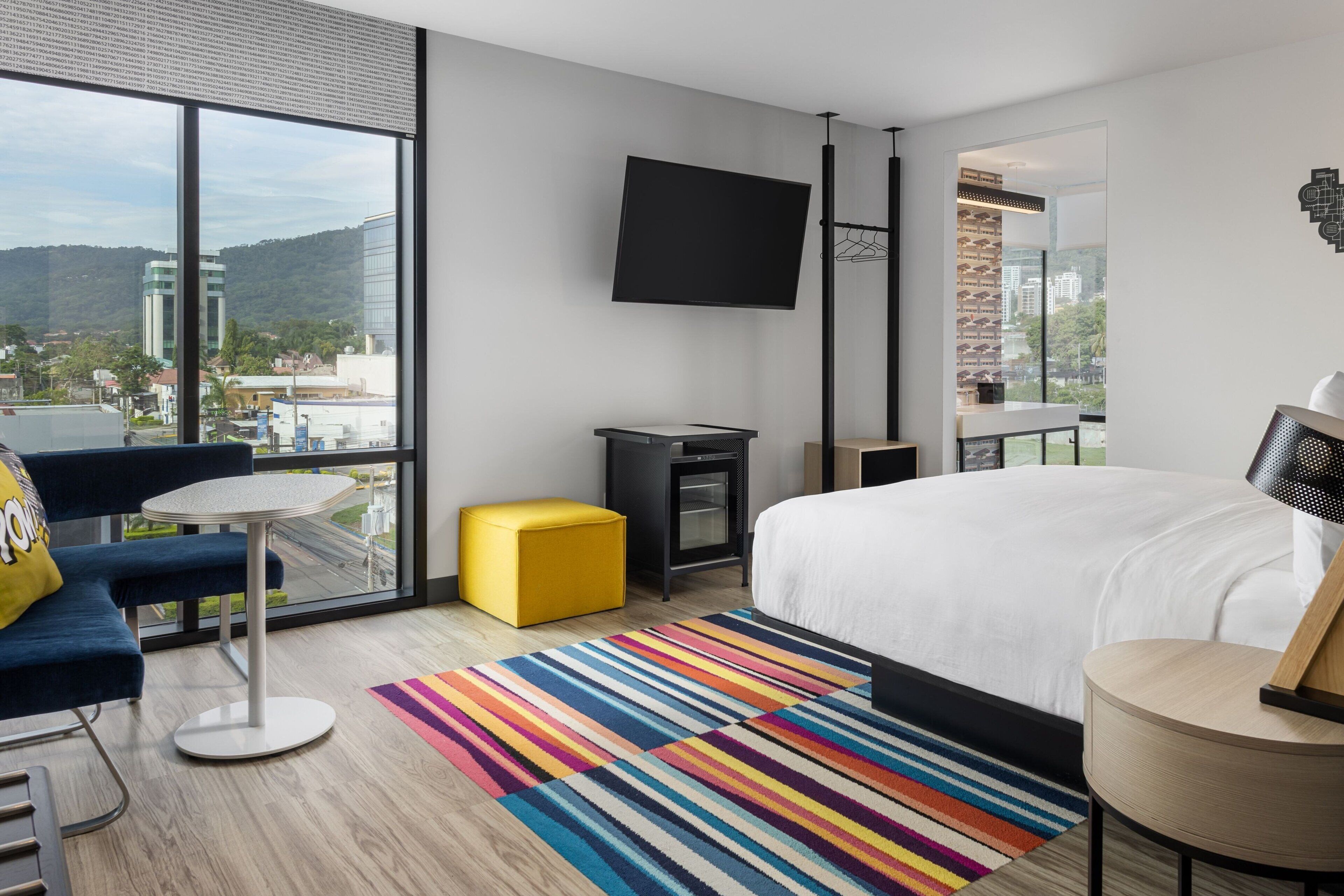 A modern hotel room with a large bed, a TV mounted on the wall, a small table, a colorful striped rug, and a view of the city through large windows.