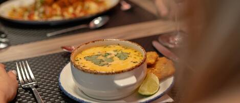 A bowl of soup on a plate with a lime wedge, accompanied by a fork and spoon on a table.