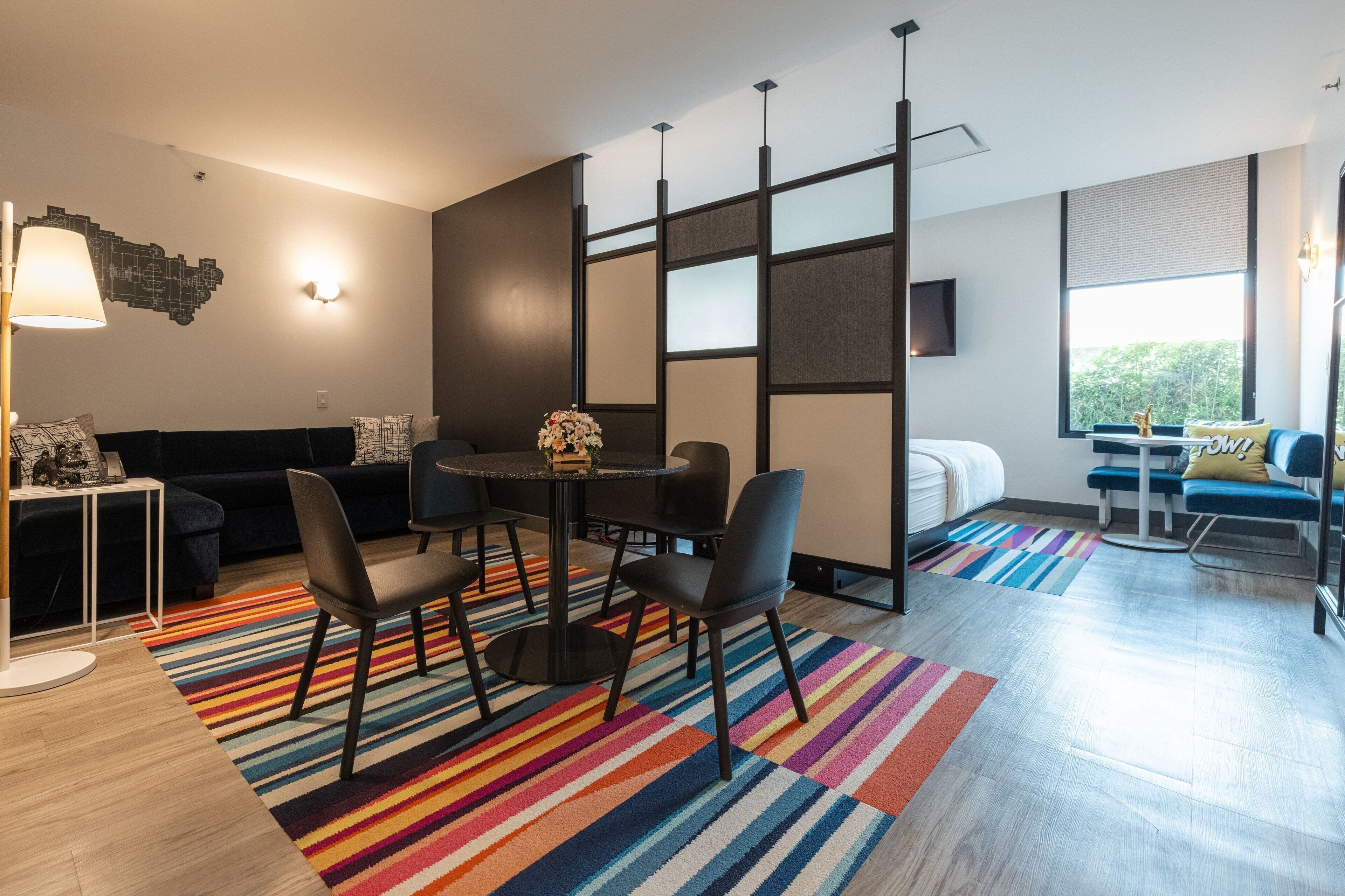 Una sala de estar moderna con un sofá oscuro, una mesa de comedor redonda con sillas, un tapete a rayas coloridos y un ventanal con vistas a la vegetación.