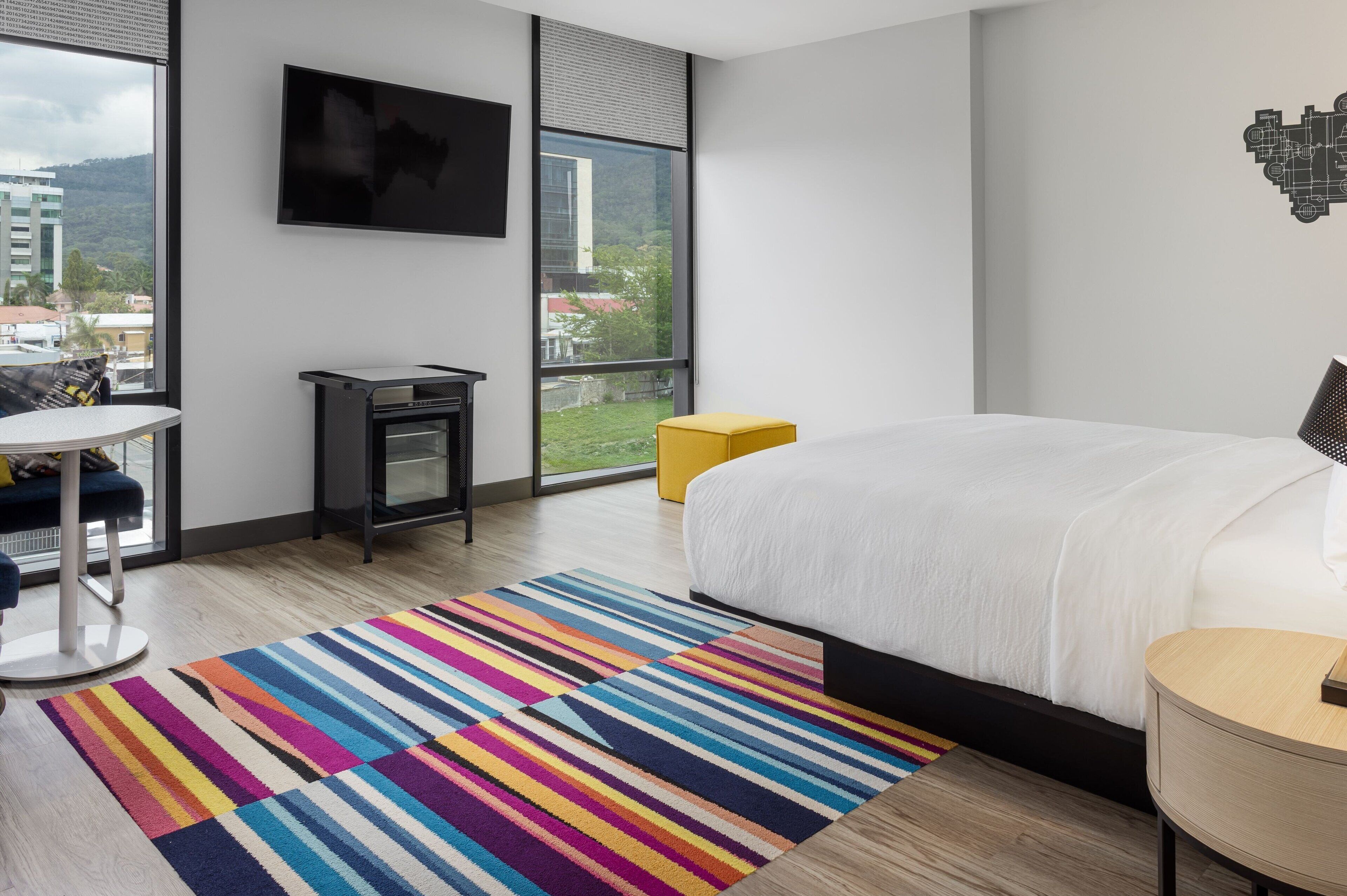 A modern hotel room with a large bed, a small desk, a chair, a colorful striped rug, and a large window offering a city view.