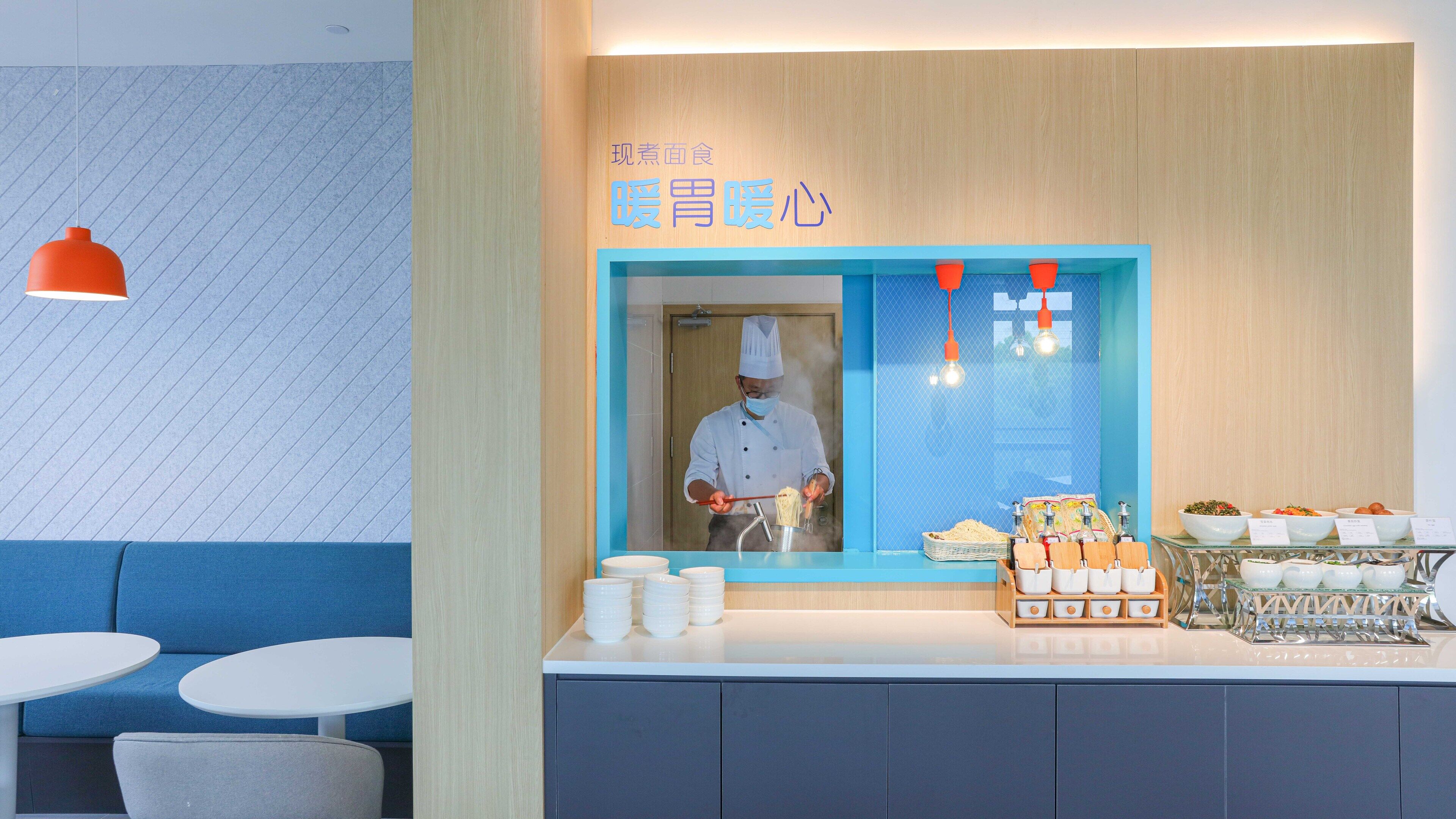 A restaurant interior with a chef preparing food, a counter with bowls of food, and seating area with tables and chairs.