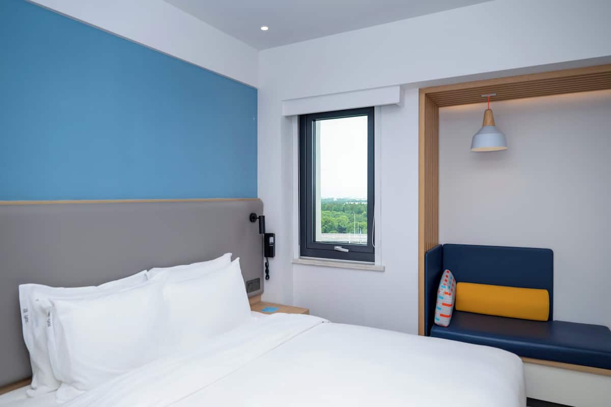 Une chambre d’hôtel comprenant un lit, une table de chevet, un téléphone, une fenêtre avec vue et un banc avec un coussin.