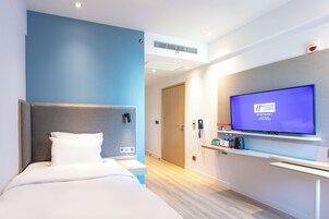 A hotel room with a bed, a television on the wall, and a small shelf with items.