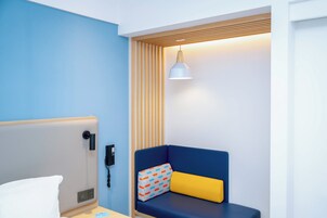 A hotel room with a bed, bedside table, a phone, a blue and yellow sofa, and a wall-mounted lamp.