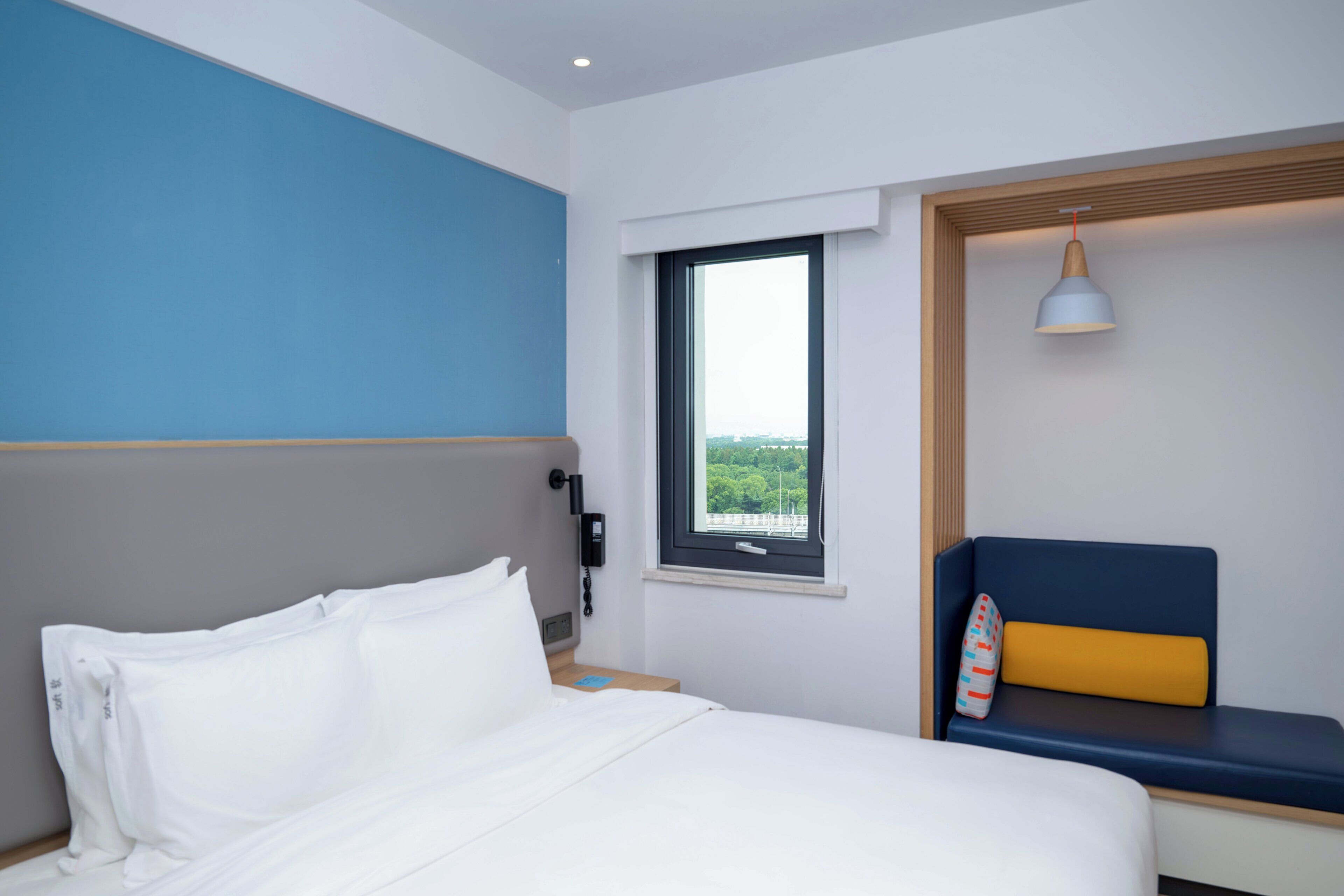 A hotel room with a bed, a window with a view, a bedside telephone, and a blue and yellow bench.
